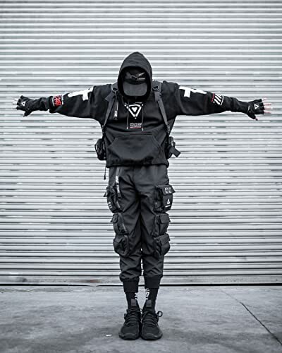 Person in black tactical streetwear with hood and mask in front of corrugated metal background.